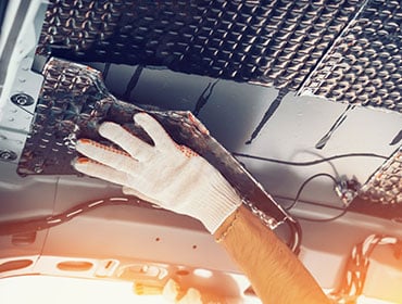 A gloved hand pressing a foil-faced barrier into place inside a vehicle bodyshell.